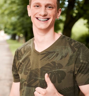 Vertical portrait of a young man, standing with thumb up in park, smiling with dental braces. Satisfied student after dentist visit. Concept of medicine and dentistry